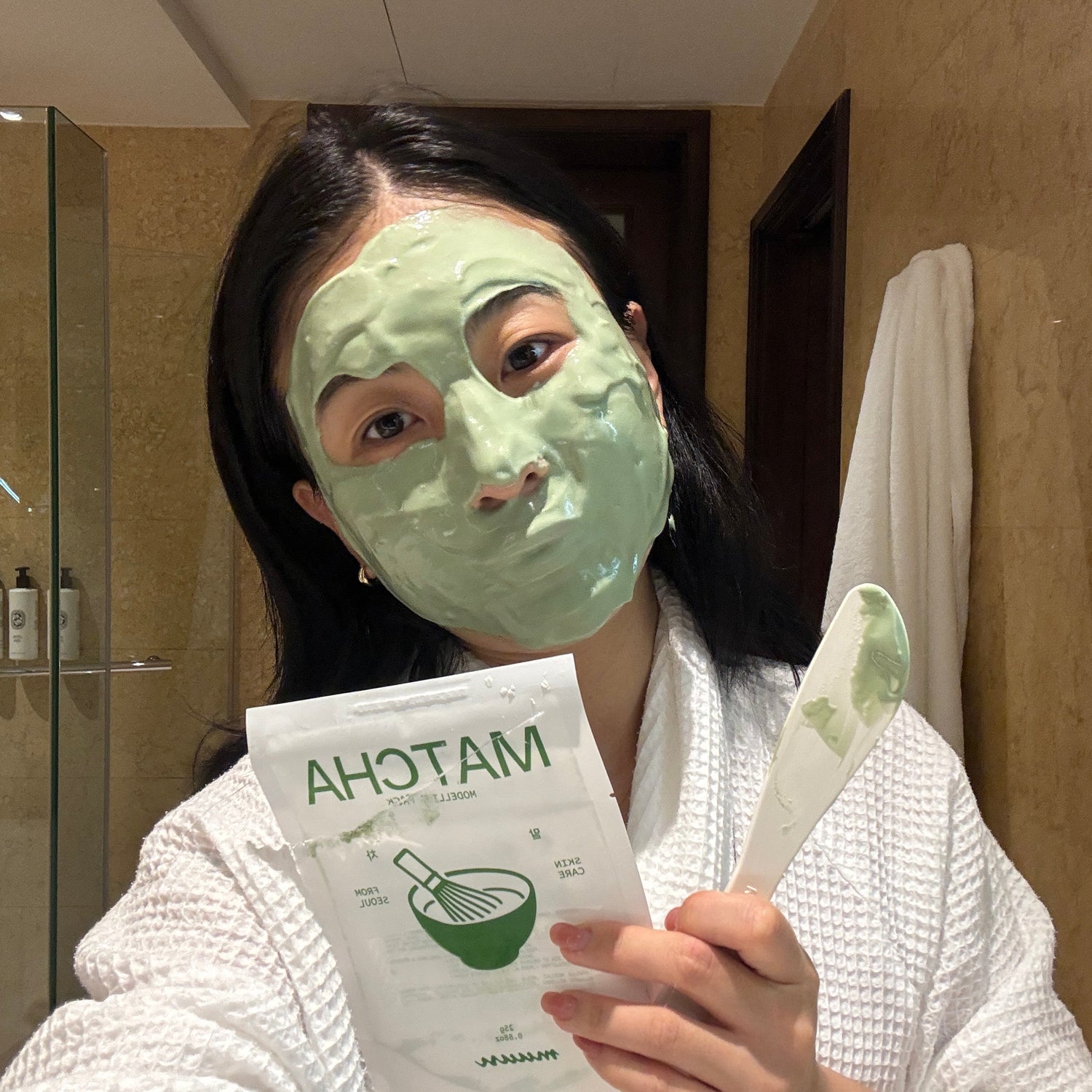 Person wearing a green facial mask and holding a box of Matcha facial masks in a bathroom.